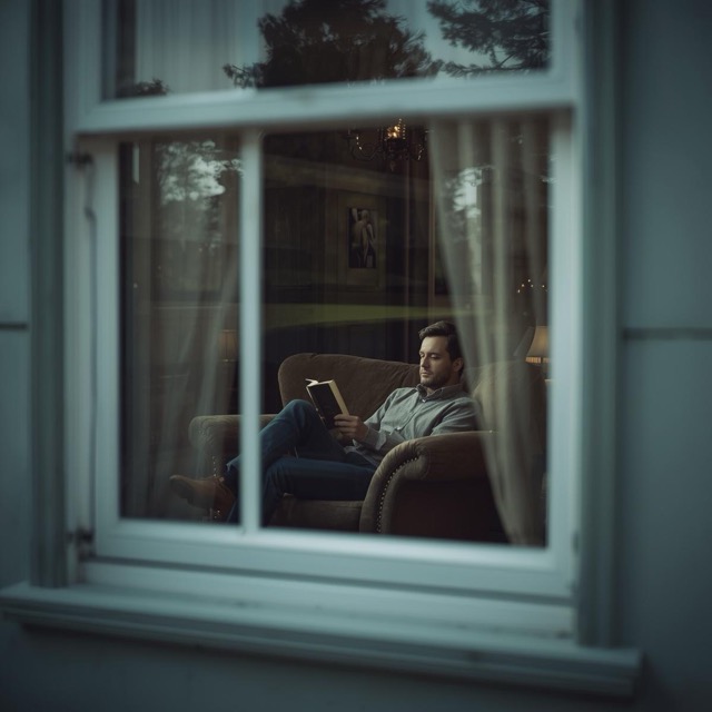 man sitting on couch reading book, looking at him through the outside window looking in (1) Medium man sitting on couch reading book